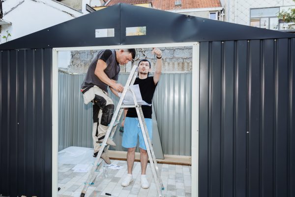 Two Caucasian men on a stepladder together talking, looking at paper instructions for assembling a metal shed on a summer day in the backyard of a house, side view, medium shot. Concept of assembling a metal shed, teamwork.